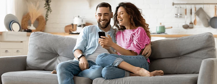 Casal jovem sentado no sofá, a sorrir, com telemóvel na mão