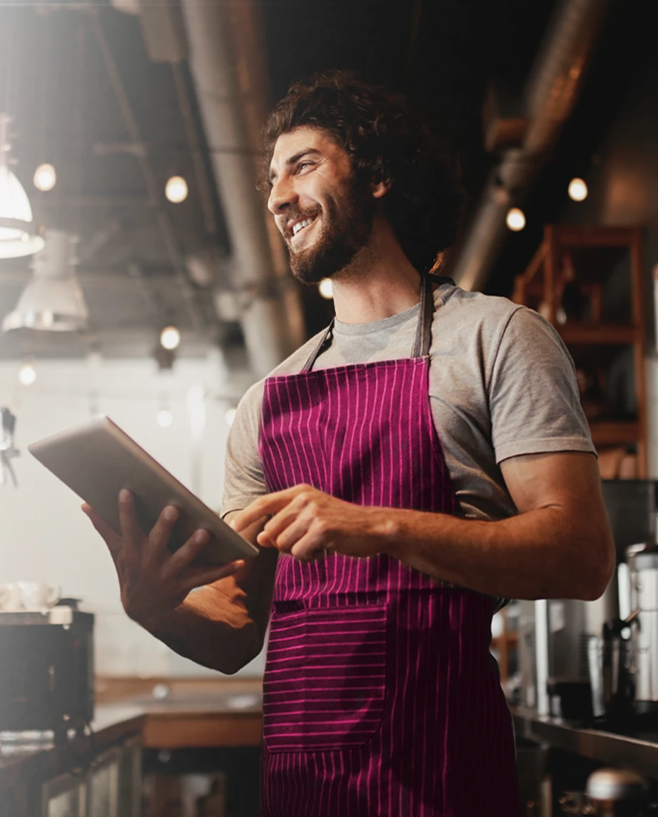 young man with a beard smiling behind the café counter, wearing an apron and holding a tablet