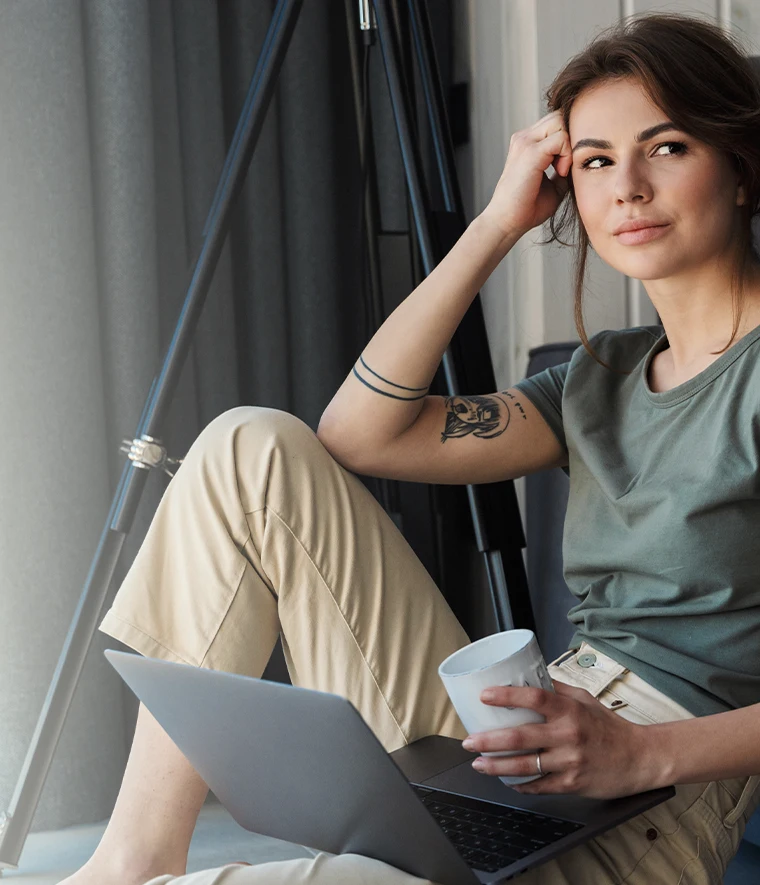 young woman with a dreamy look, sitting on the floor with a laptop and a coffee cup