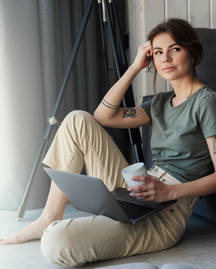 mulher jovem com ar sonhador, sentada no chão com laptop e copo de café