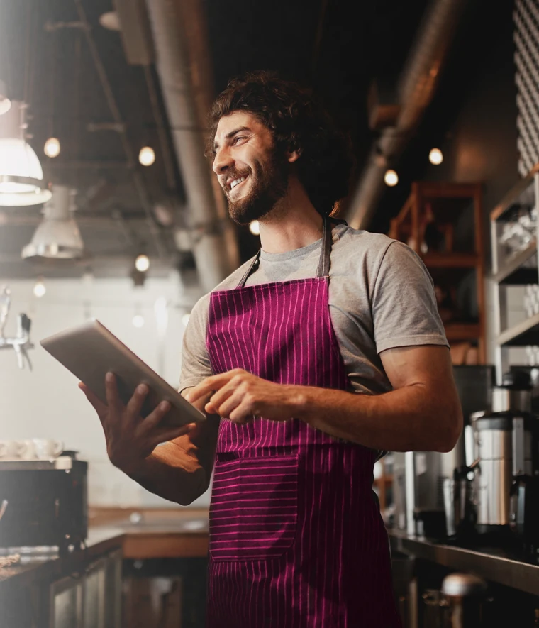 young man with a beard smiling behind the café counter, wearing an apron and holding a tablet