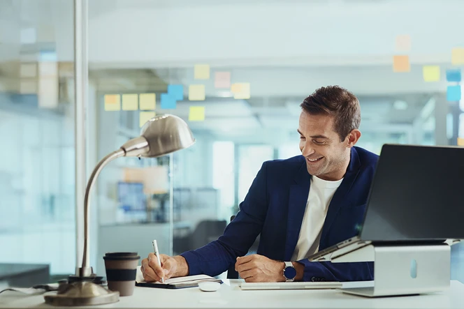 Smiling man behind a desk, writing in a notebook, with a computer beside him
