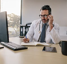 médico a atender paciente por telemóvel, a escrever num caderno e com computador em frente