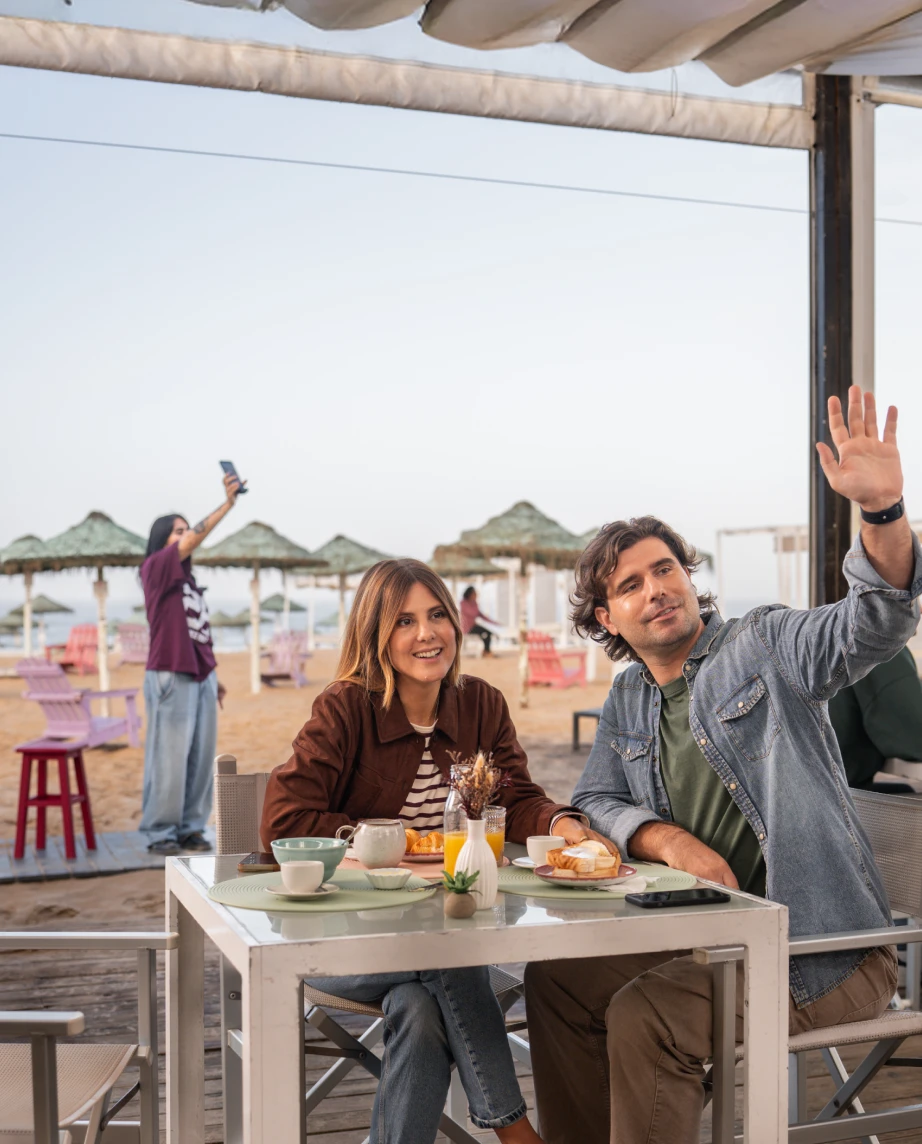 Um homem e uma mulher sentados num bar de praia, e o homem a acenar a alguém