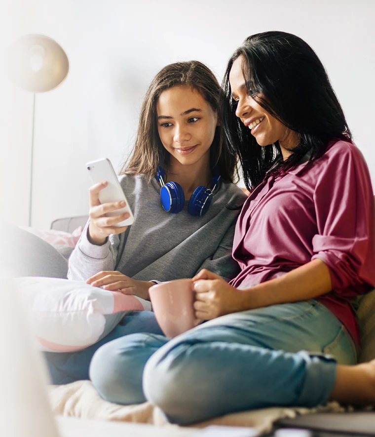mother holding a cup of tea and the teenage daughter with a phone, sitting on the sofa in a moment of closeness