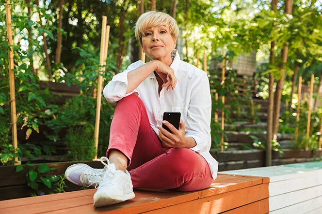 Senhora de meia idade sentada em banco de jardim  com as pernas cruzadas e com o telemóvel na mão