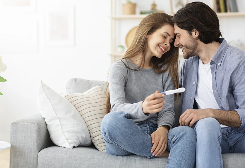 casal sentado no sofá, feliz, ela a segurar um teste de gravidez
