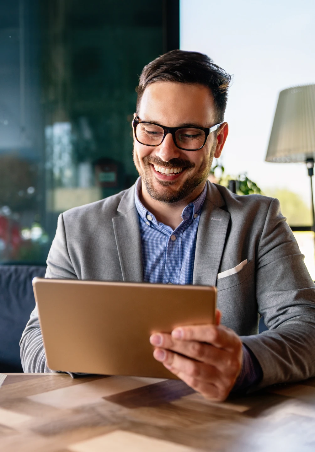 Homem com óculos pretos, camisa azul e branca aos xadrez e blazer cinza, a sorrir para o tablet que tem na mão direita