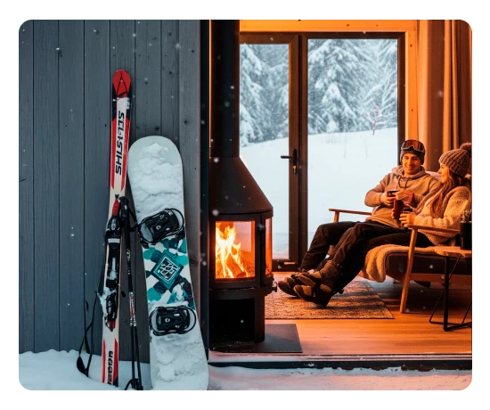 Esquis e snowboard encostados junto a uma cabana na neve; casal sentado no interior aquece-se junto à lareira após atividade de inverno