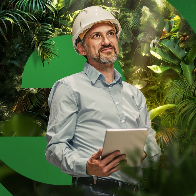 Homem, com uma camisa azul clara e um capacete de segurança branco, a segurar um tablet nas duas mãos, à frente de um fundo de cor verde e conjuntos de plantas