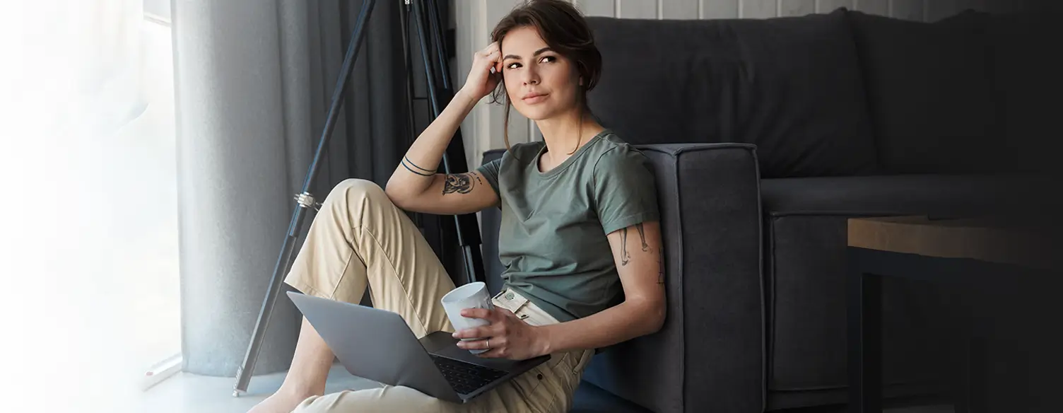 young woman with a dreamy look, sitting on the floor with a laptop and a coffee cup