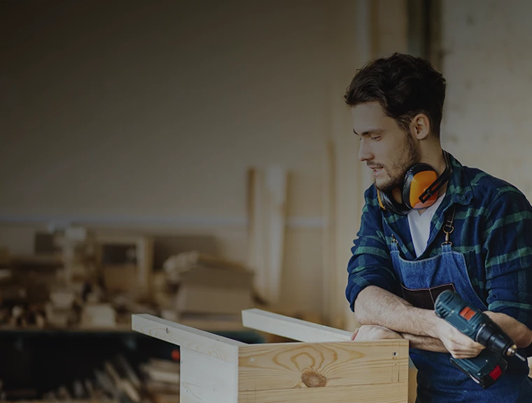 Jovem rapaz serralheiro a construir uma cadeira de madeira, na oficina, vestido de fato macaco de ganga e com perfuradora de bateria na mão