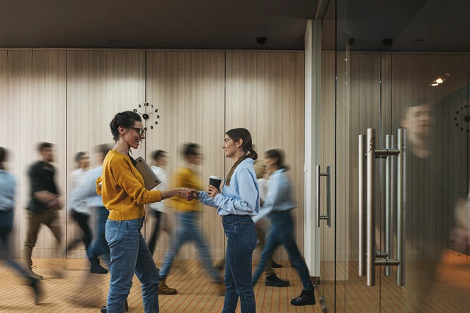 2 women shaking hands in a company hallway with several people walking by