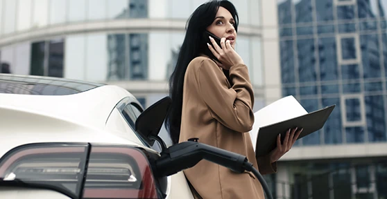 senhora a falar ao telemóvel e com caderno na mão, encostada a carro elétrico em carregamento