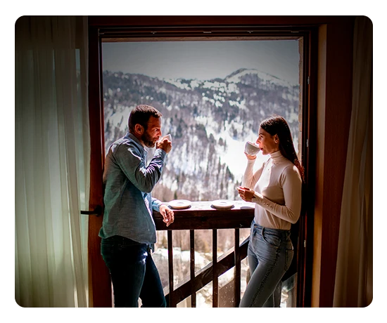 Casal a beber uma bebida quente no parapeito de uma janela, com uma montanha com neve na paisagem