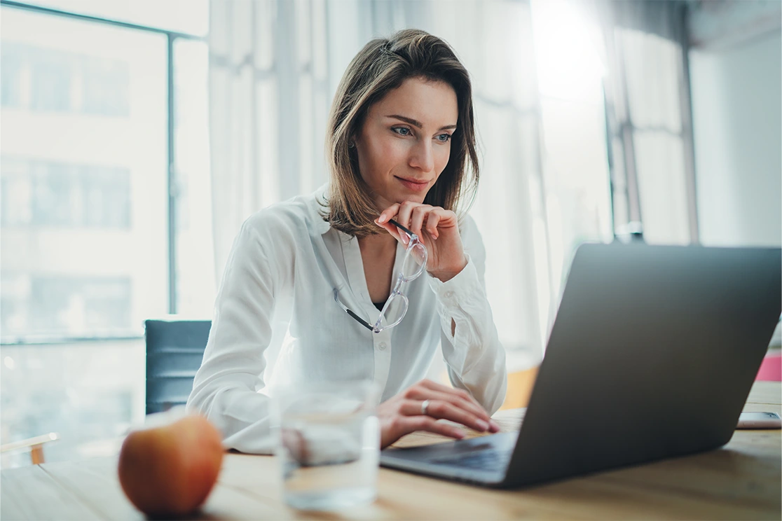 woman sitting on desk, holding glasses, in front of pc