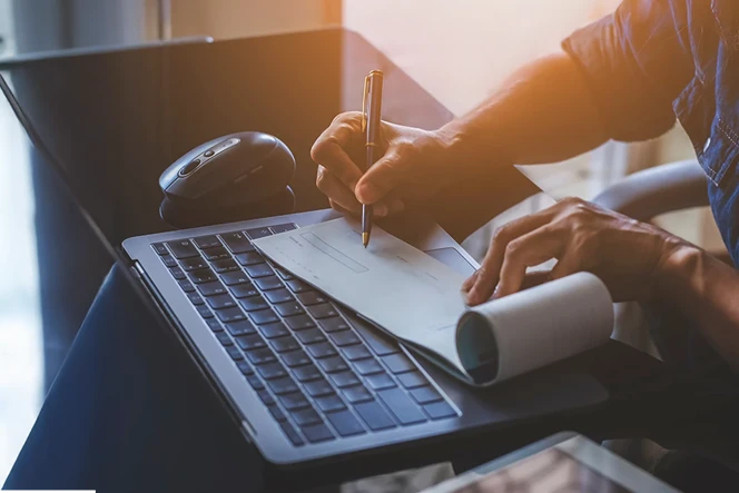 Close-up of hands signing a check on a desk with a computer, tablet, and pen, under sunset lighting