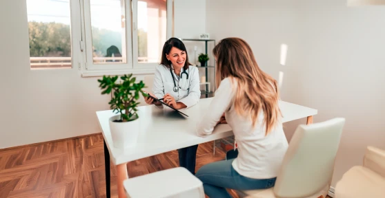Médica a sorrir, sentada numa secretária a atender uma uma paciente