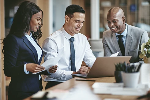 2 homens e 1 mulher, sorridentes, numa sala de reuniões com uma secretária com portátil e papéis
