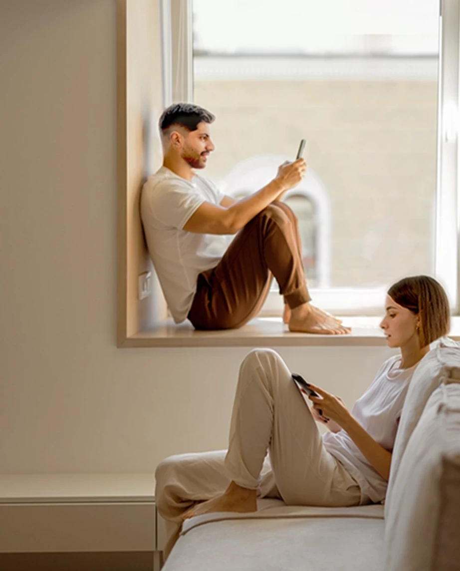 Casal jovem a consultar telemóvel. Ele sentado na janela e ela no sofá