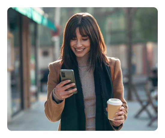 Mulher sorridente na rua a olhar para o telemóvel enquanto segura um copo de café