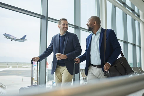 Dois homens no Aeroporto, a caminhar lado a lado a conversar, transportando a suas bagagens