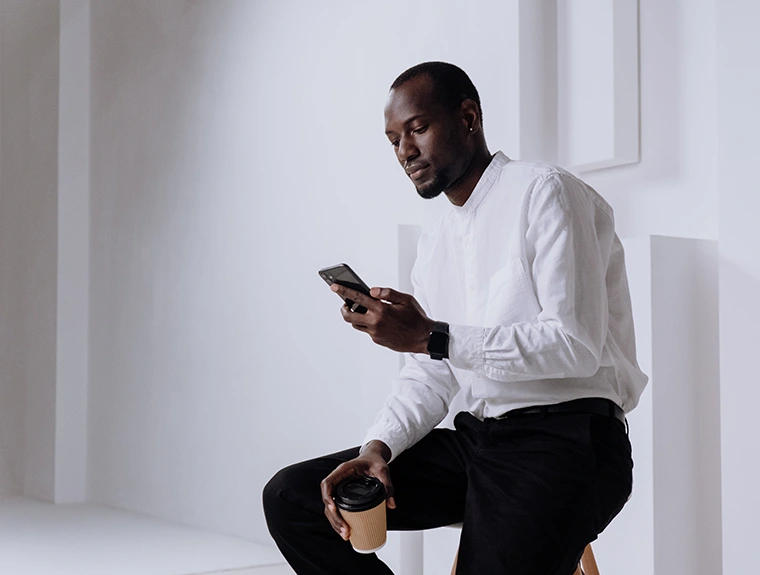 homem com camisa branca e calças pretas, sentado num banco, com telemóvel numa mão e copo de café na outra
