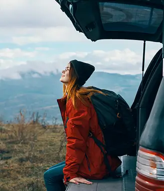 mulher sentada na bagageira do carro, a olhar para paisagem com montanhas