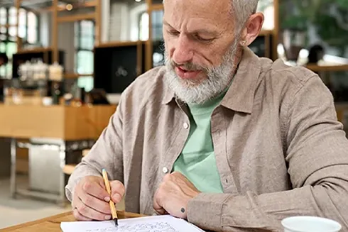 Senhor sentado à mesa, a tomar notas num caderno e com chávena em cima da mesa
