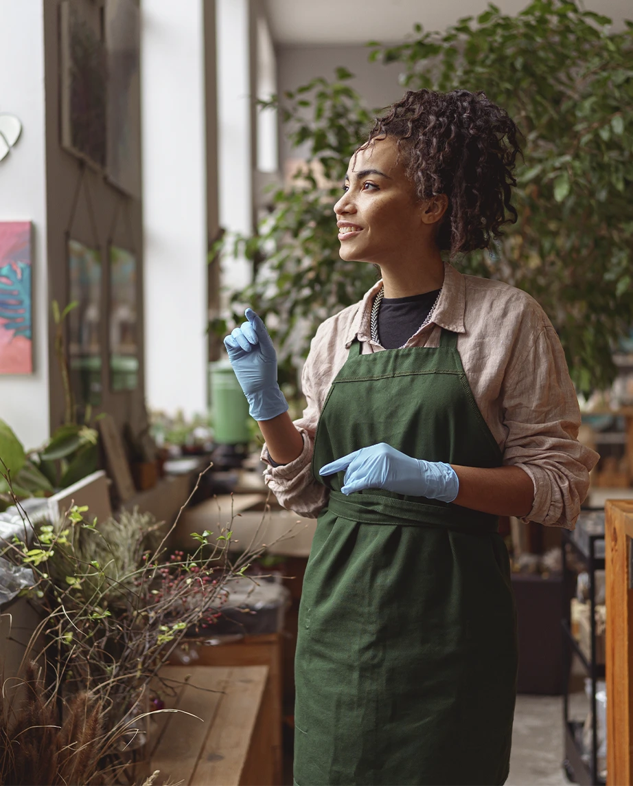 Senhora  num espaço tipo estufa com uma luvas azuis e um avental a olhar pela janela e a sorrir