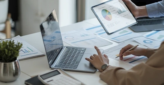 Duas pessoas a analisar gráficos num portátil e num tablet sobre uma mesa com documentos