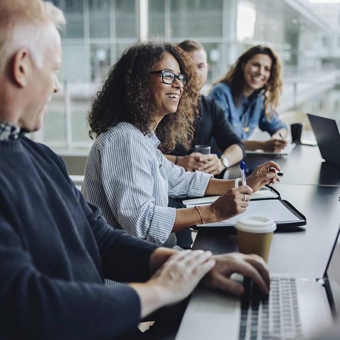 Uma mulher sorridente com óculos e cabelo encaracolado está sentada numa mesa de reunião com colegas de trabalho.