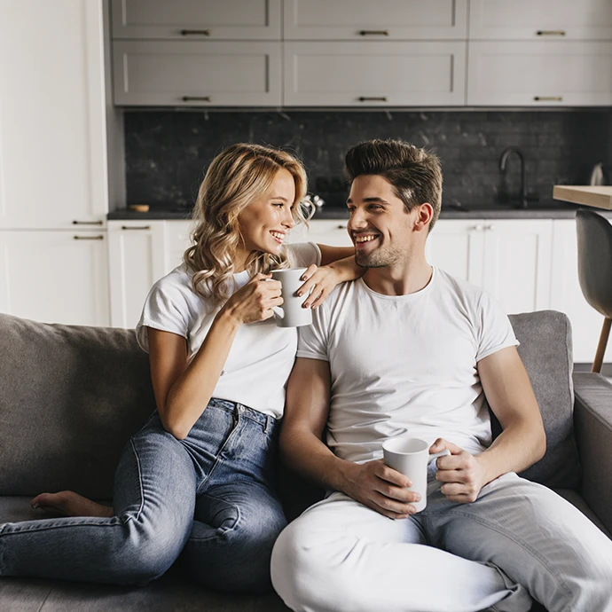 jovem casal, sentados no sofá encostados um ao outro, com chávenas na mão