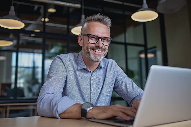 smiling man with a beard and glasses, sitting at a desk with an open laptop