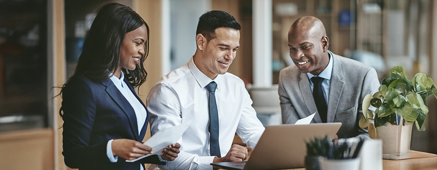 2 homens e 1 mulher, sorridentes, numa sala de reuniões com uma secretária com portátil e papéis