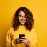 mulher, vestida de amarelo e com parede de fundo amarela, sorridente e com telemóvel na mão