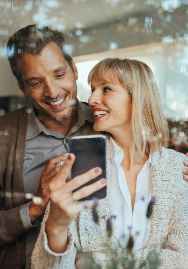 Casal sorridente a olhar para um telemóvel juntos, refletidos numa janela