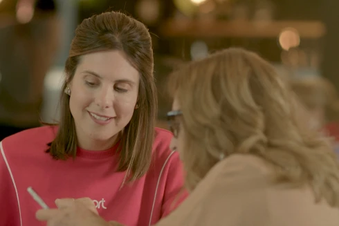 Rapariga na casa dos 30 anos com uma camisola cor-de-rosa e uma senhora mais velha a mostrar-lhe o seu telemóvel