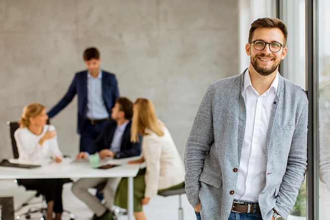 Smiling man leaning against a glass window in a meeting room, with four people in the background