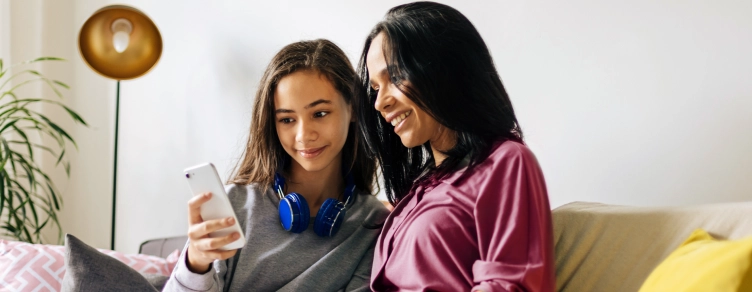 mother holding a cup of tea and the teenage daughter with a phone, sitting on the sofa in a moment of closeness