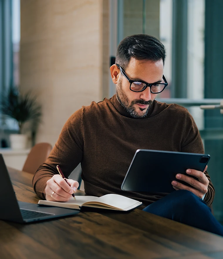 man sitting at the desk, writing notes and consulting a tablet