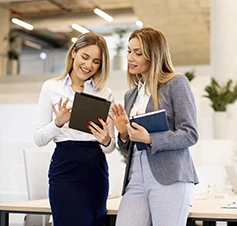 2 mulheres numa sala de reuniões a conversar, com 3 pessoas ao fundo