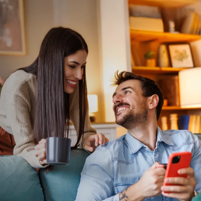 Casal em casa, sorridente no sofá; ele usa o telemóvel enquanto ela segura uma chávena de café