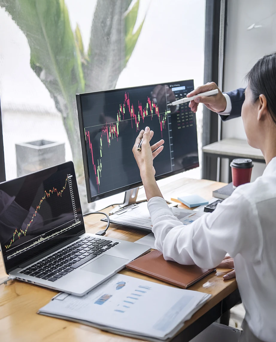 2 pessoas num escritório, com 2 computadores com gráficos, a analisar mercados