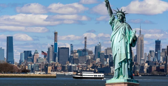 Vista panorâmica de Nova Iorque com a Estátua da Liberdade em primeiro plano à direita e os arranha-céus de Manhattan ao fundo sob um céu azul.