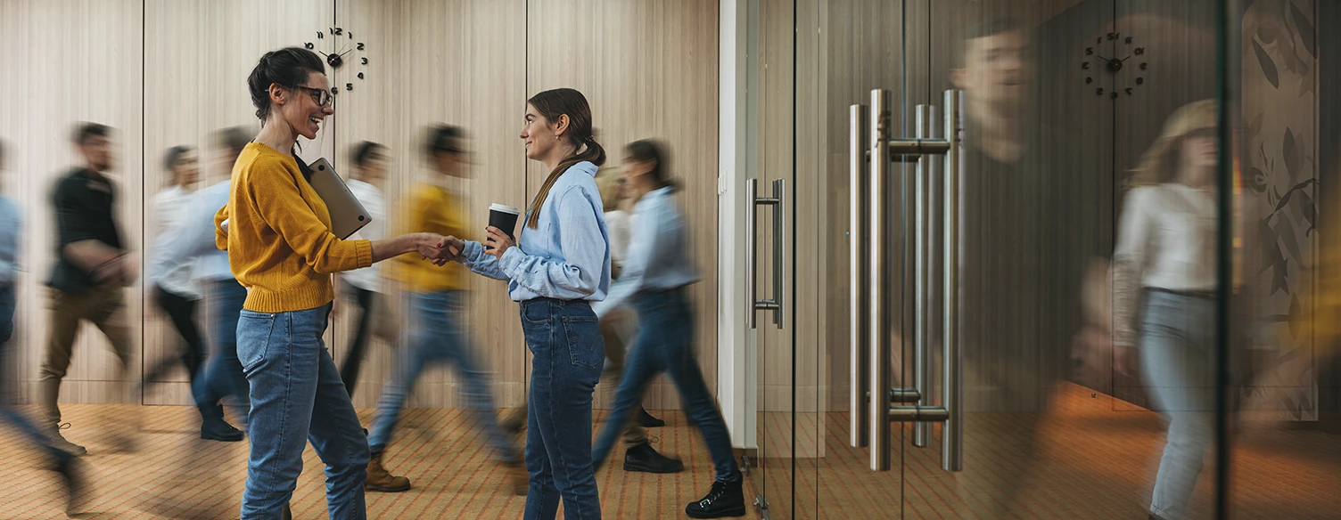 2 mulheres num aperto de mão, num corredor de empresa com várias pessoas a passar