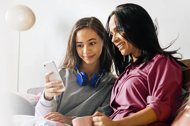 mother holding a cup of tea and the teenage daughter with a phone, sitting on the sofa in a moment of closeness