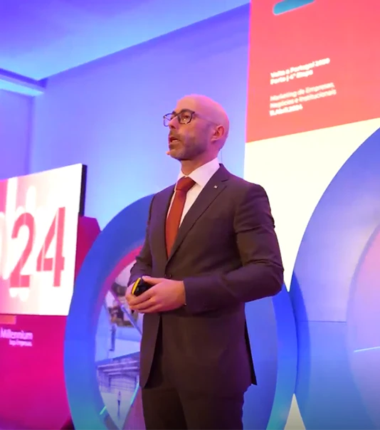 Homem, de fato e gravata verde, a dar uma palestra e com um powerpoint atrás com o headline "Juntos Lideramos o Futuro"