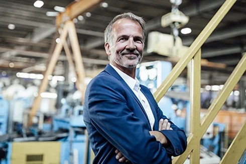 homem vestido de fato azul, com braços cruzados e sorridente, numa fábrica