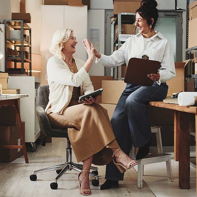 Duas senhoras a bater as mãos uma na outra e a sorriem no local de trabalho. Uma delas está sentada em cadeira e a outra encostada na mesa